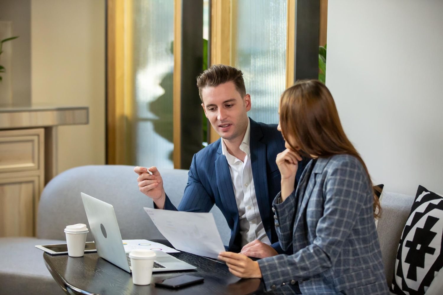 Two coworkers reviewing documents on a sofa beside a laptop and coffee mugs