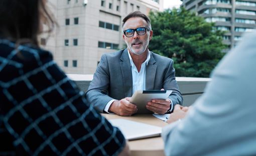 Business meeting outdoors, man in gray blazer holding a tablet across a table.