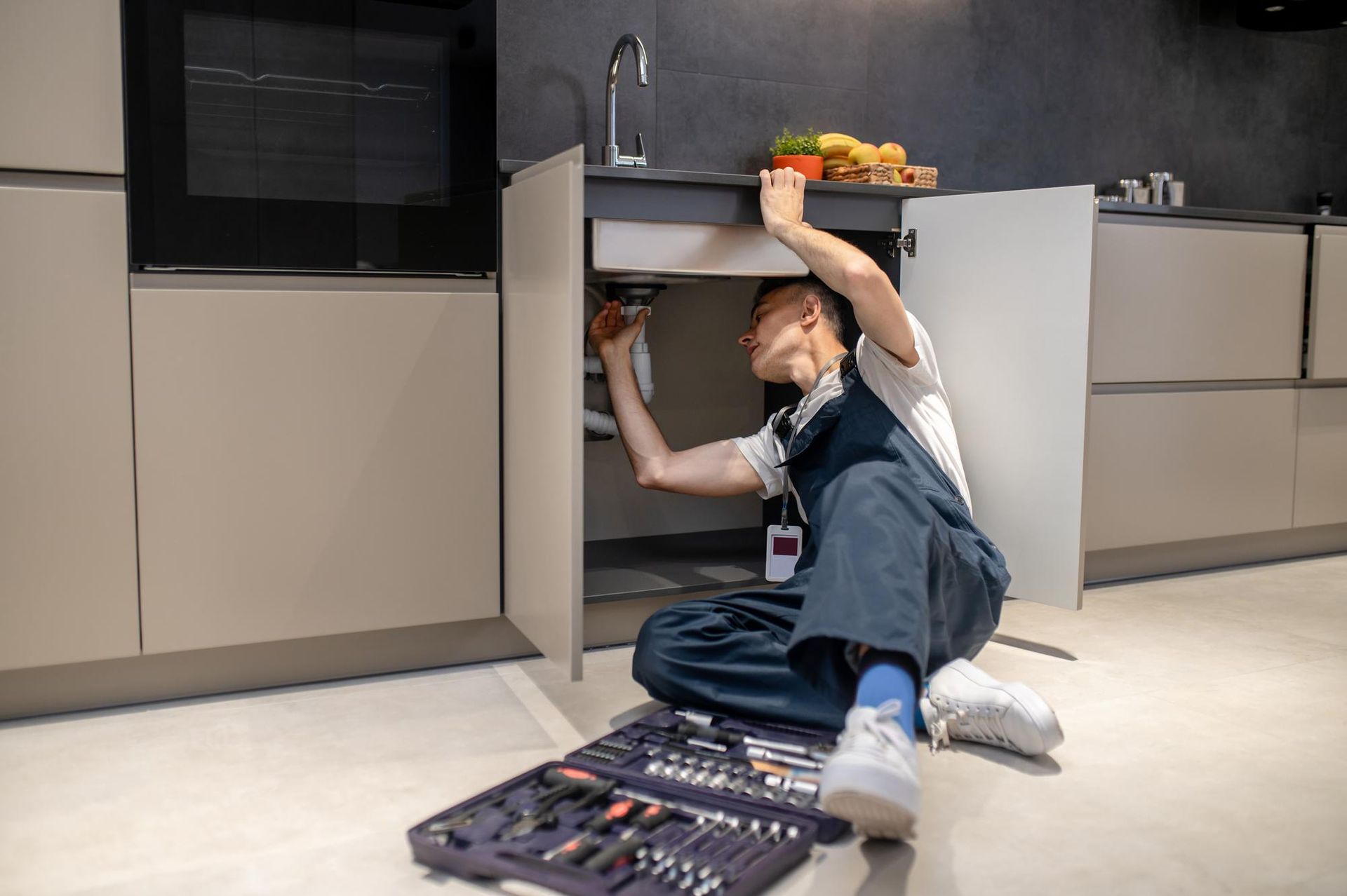Person lying under a kitchen cabinet reaching upward, with tools on the floor and fruit on the counter.