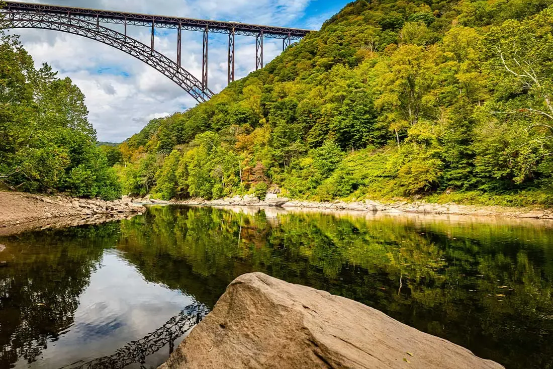 Forest river beneath a tall steel arch bridge reflected in calm water, with rocky shore and green hills