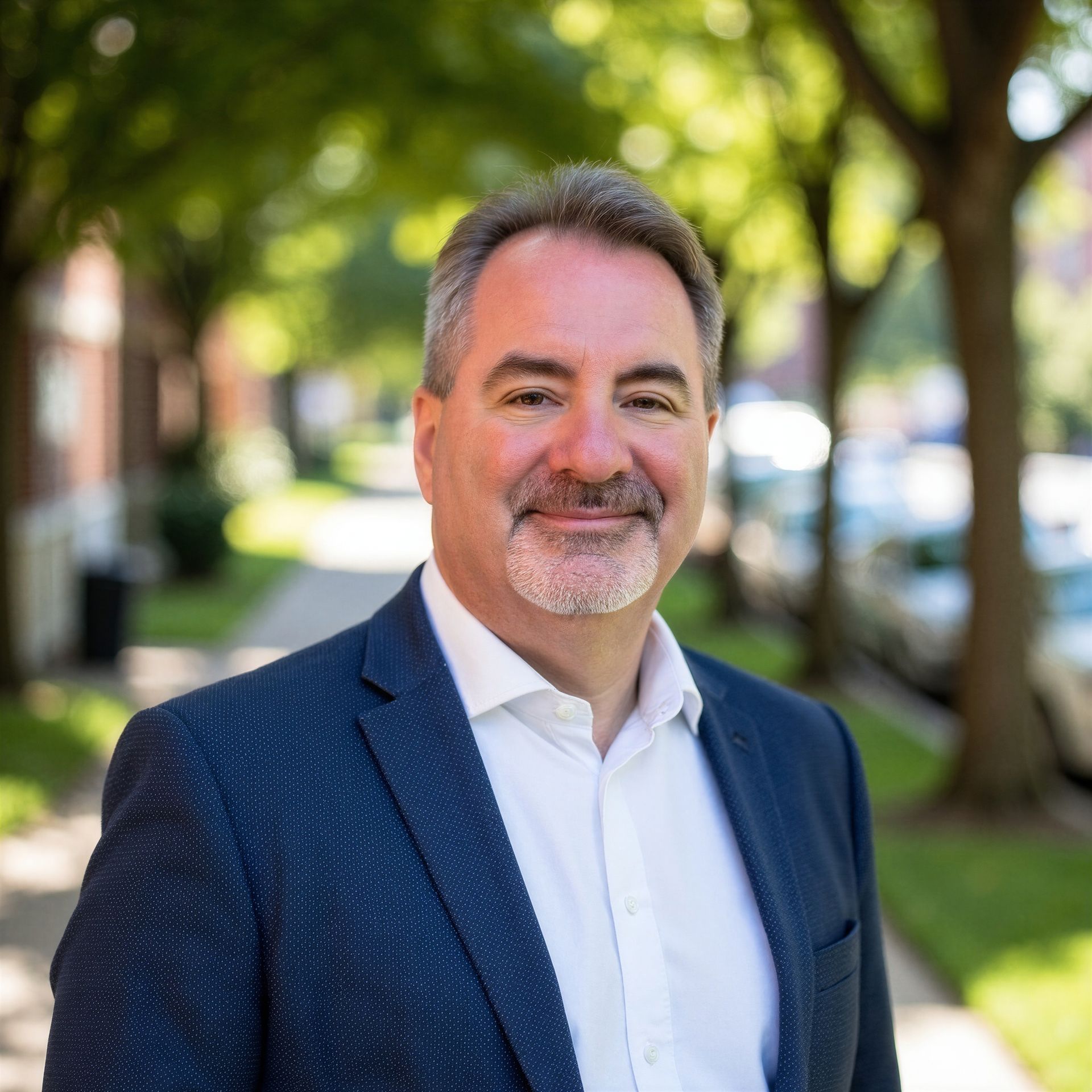 Portrait of a smiling man in a navy blazer and white shirt outdoors on a tree-lined street