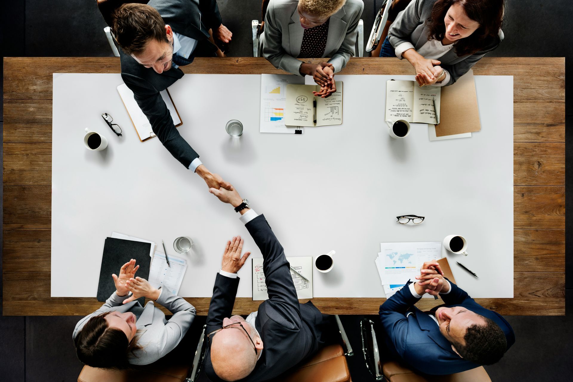 Colleagues in a top-down office meeting, shaking hands across a white conference table with papers and coffee cups