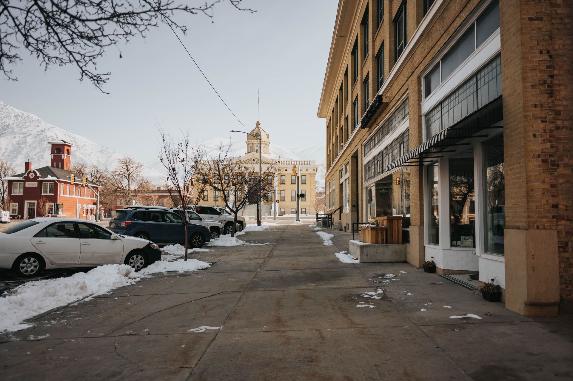 Snowy city sidewalk beside parked cars and brick buildings under a pale sky.