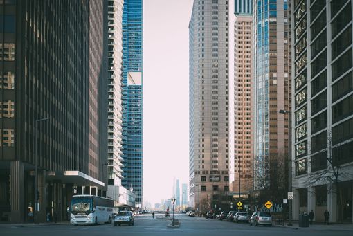 Tall downtown street framed by high-rise buildings with cars driving under a pale sky.