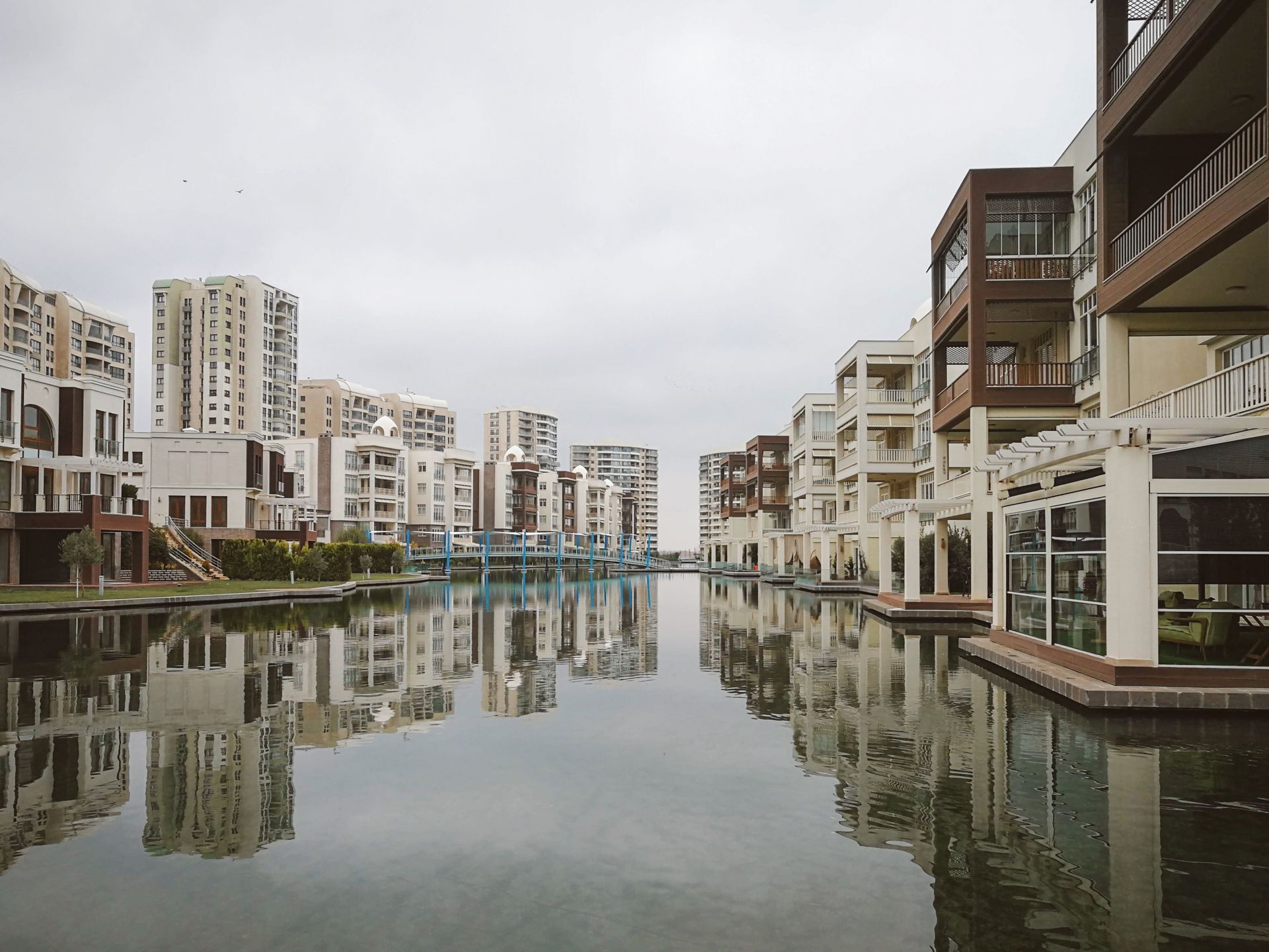 Modern waterfront canal lined with white and beige buildings, reflected in calm water under a cloudy sky