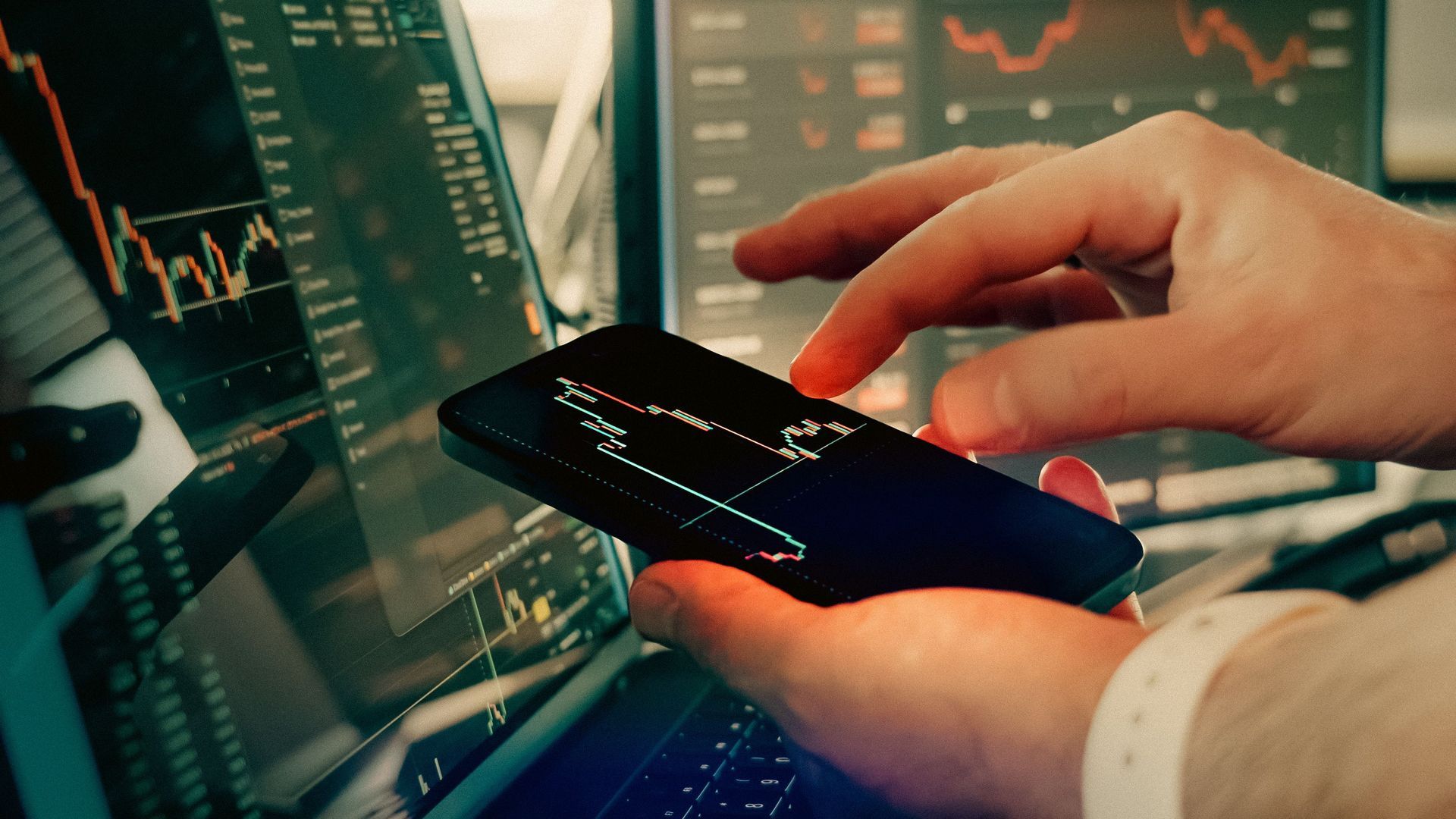 Hands holding a smartphone in front of multiple stock market charts on monitors, suggesting trading analysis