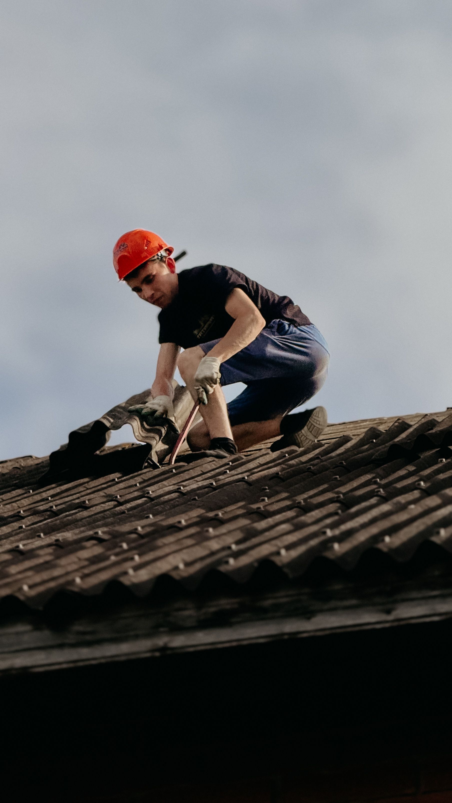 Worker in a red hard hat crouches on a tiled roof, inspecting or repairing it.