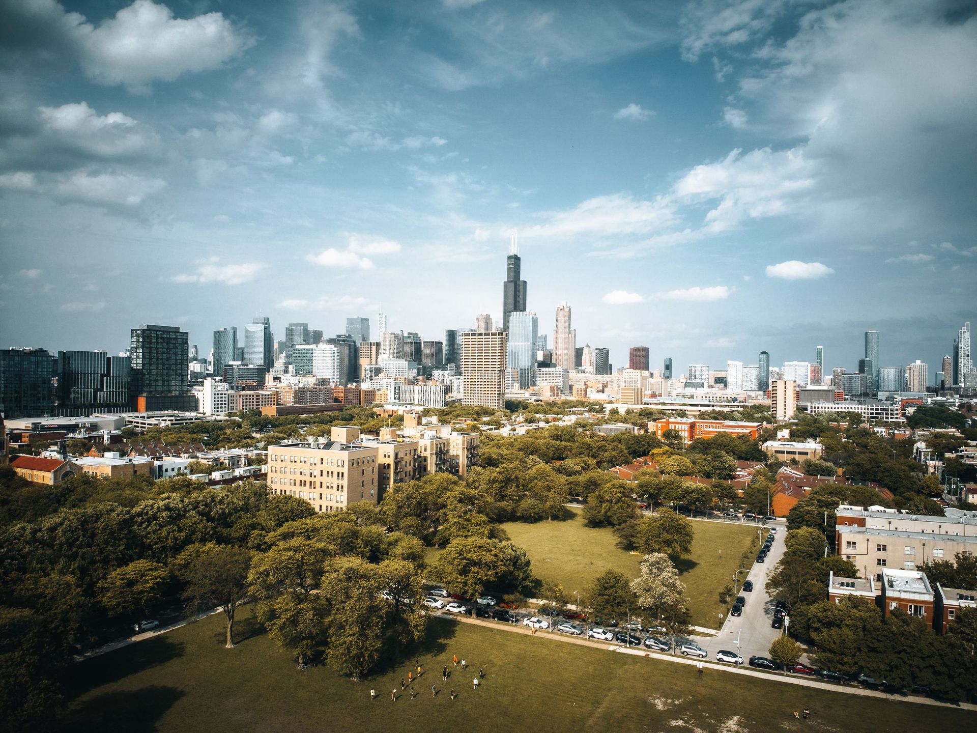 City skyline above a large green park under a bright blue, partly cloudy sky