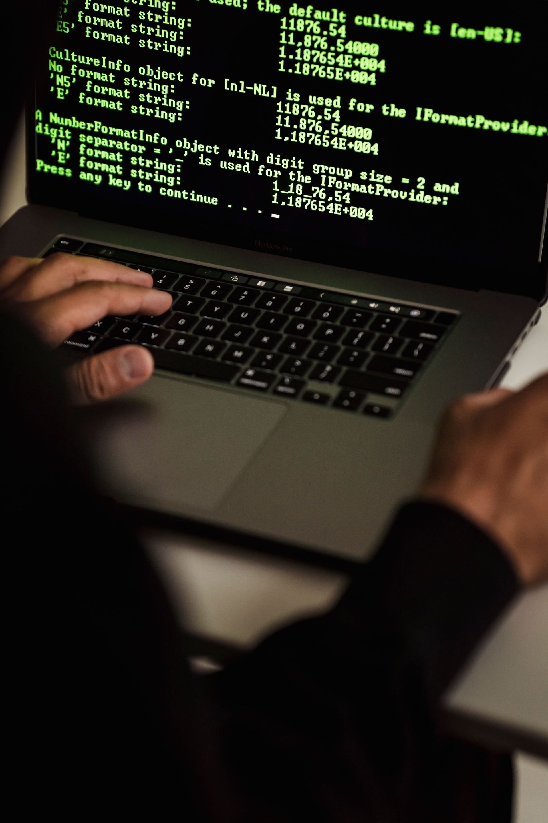 Hands typing on a laptop with green code on the screen in a dimly lit room
