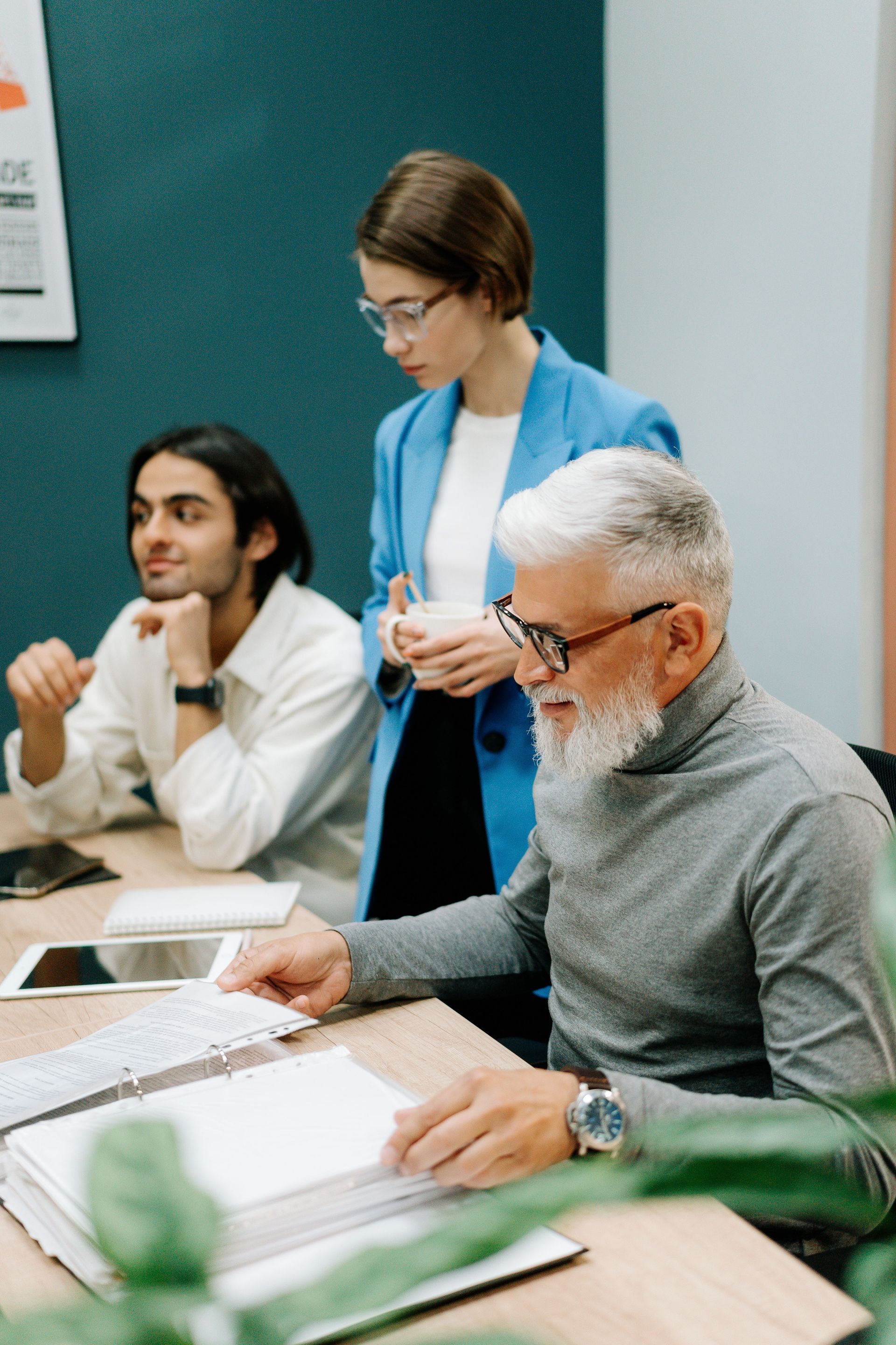 Three people in a meeting room reviewing documents at a table, one standing and two seated.