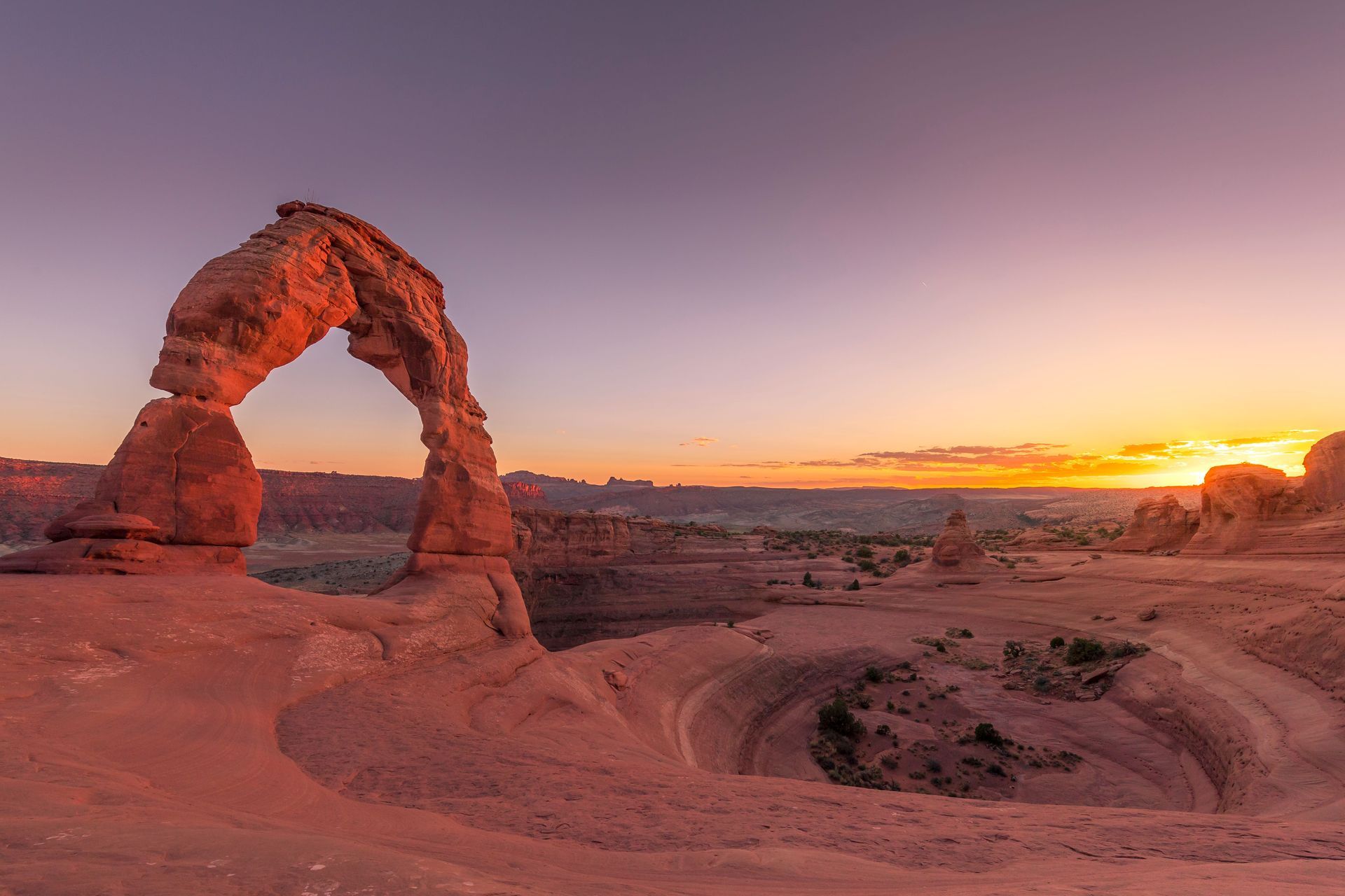 Sunset over a red sandstone arch and desert canyon, with warm orange and purple sky
