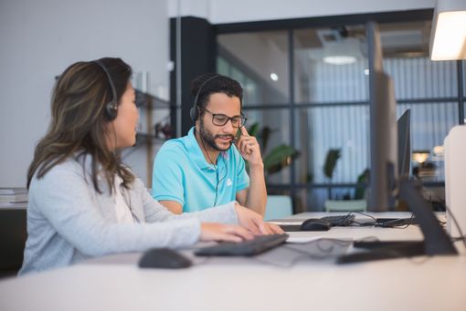 Two coworkers in an office, one on the phone and one using a laptop at a desk.