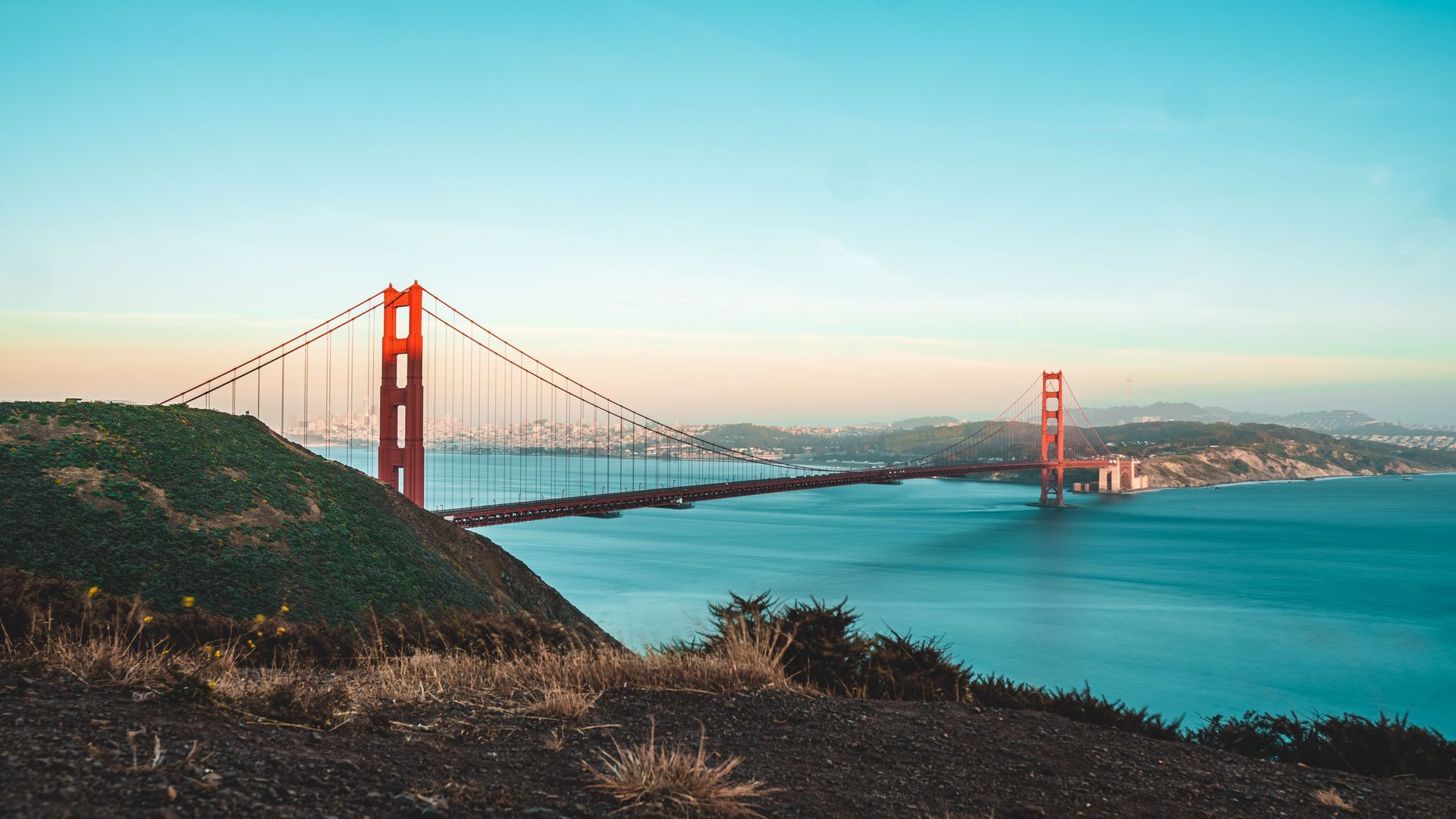 Golden Gate Bridge at sunset over blue water, viewed from a grassy hillside.