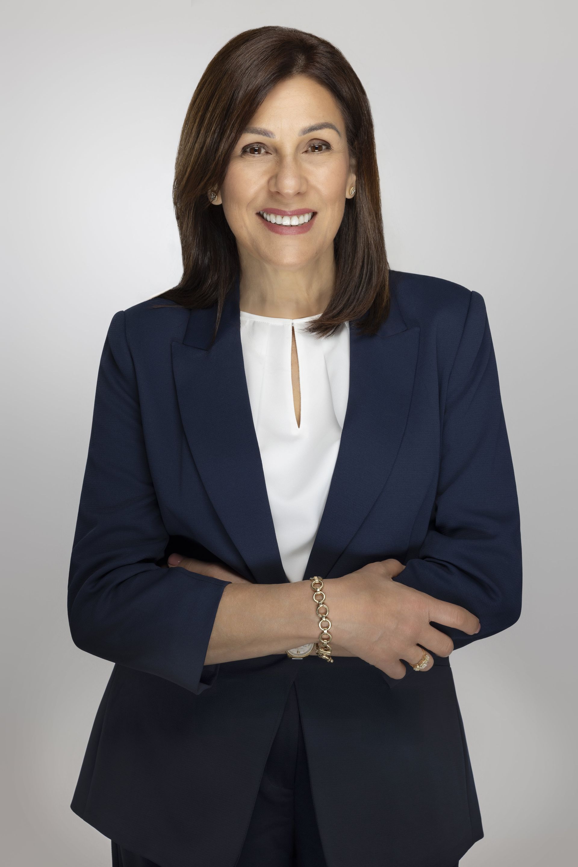 Woman in a blue blazer and white blouse, arms crossed, smiling against a gray backdrop.