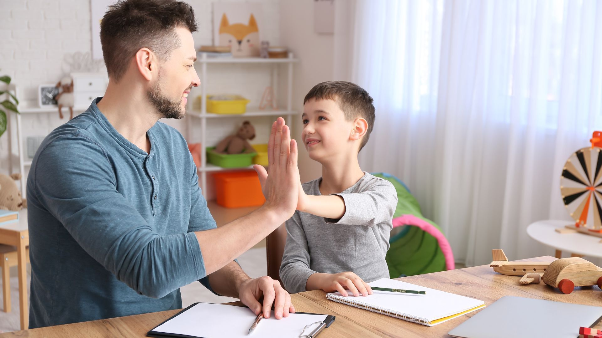 Man and boy high-five at a desk with papers. Smiling, bright room with toys.
