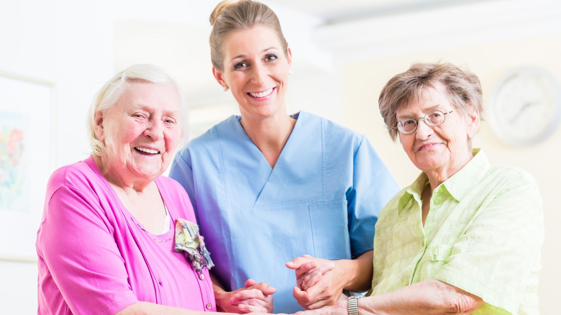 Woman in blue scrubs smiles between two older women, all smiling in a care setting.