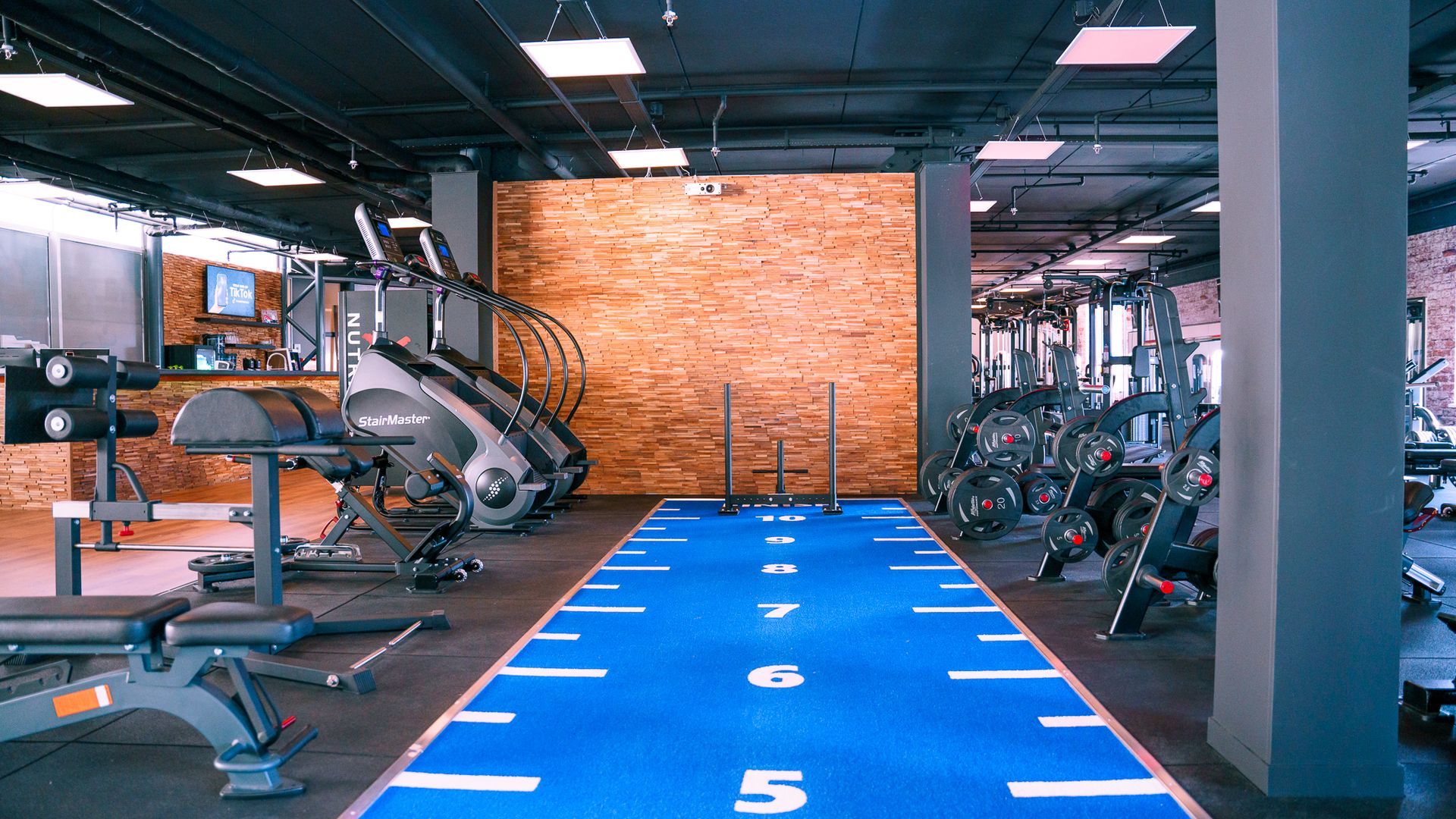 Interieur van een sportschool met een blauwe atletiekbaan, fitnessapparatuur en een muur van zichtbaar baksteen.
