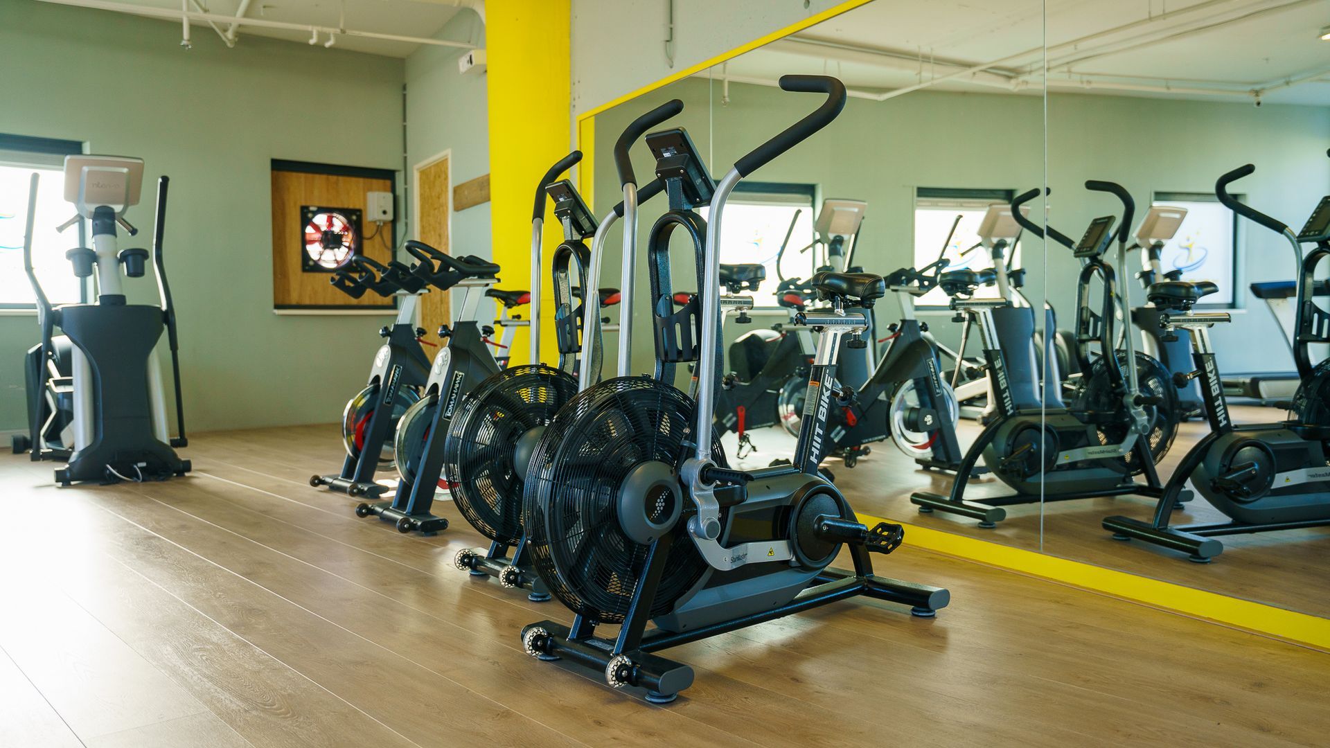 Een fitnessruimte met meerdere crosstrainers, een dartbord en een grote spiegel.