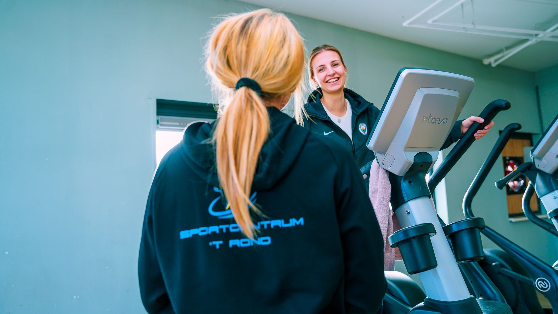 Een vrouw in een zwarte hoodie met een paardenstaart praat in de sportschool met een andere vrouw.