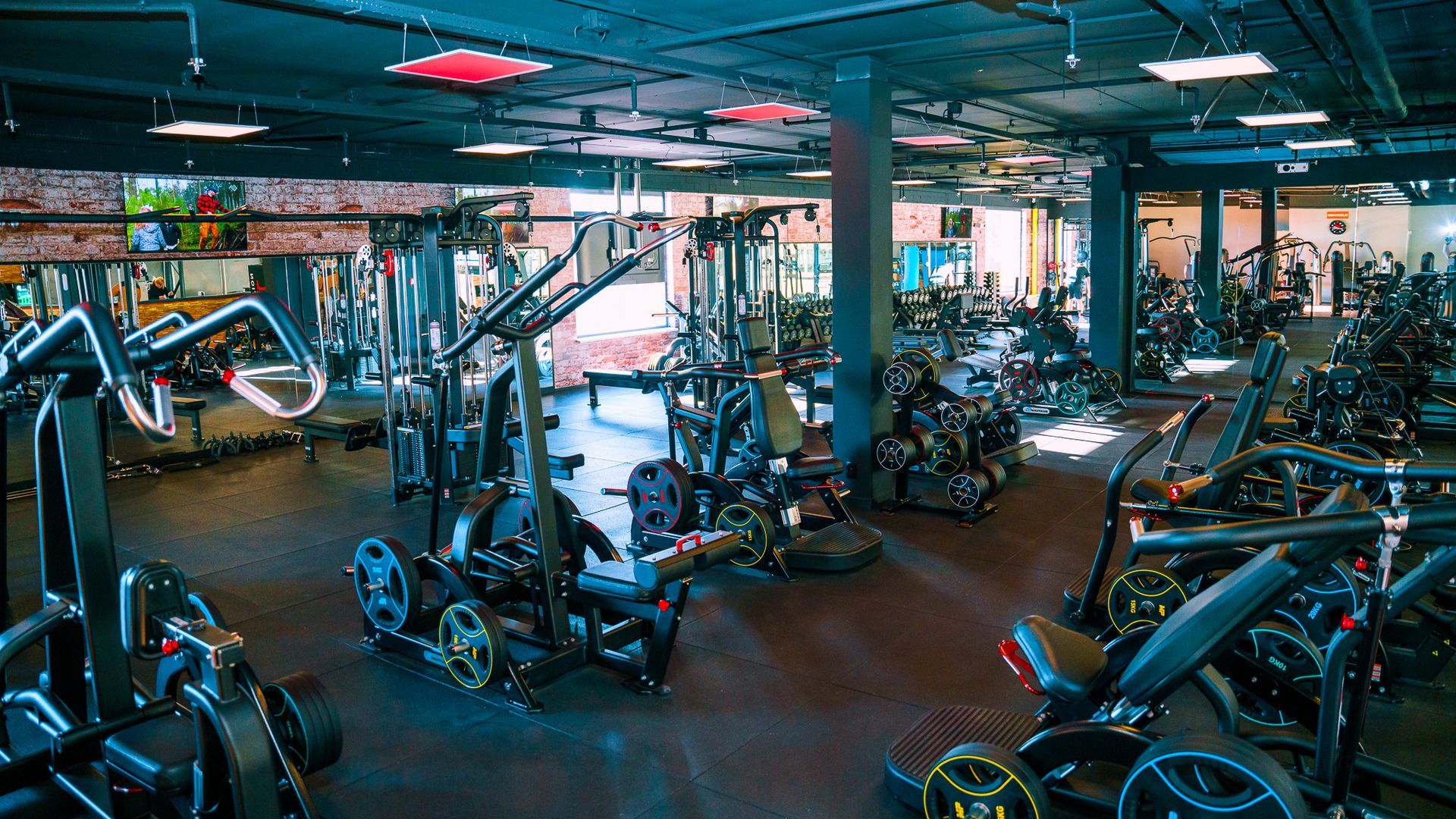 Interieur van een sportschool met diverse fitnessapparaten, donkere muren en fel licht aan het plafond.
