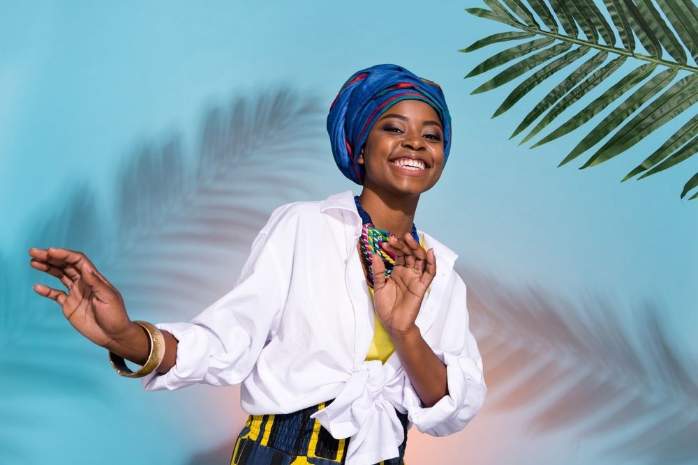 A woman wearing a turban and a white shirt is standing in front of a palm tree.
