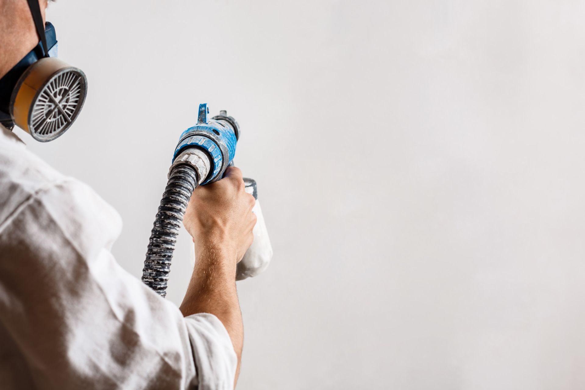 Person in white shirt and respirator spraying a white wall with a paint sprayer.