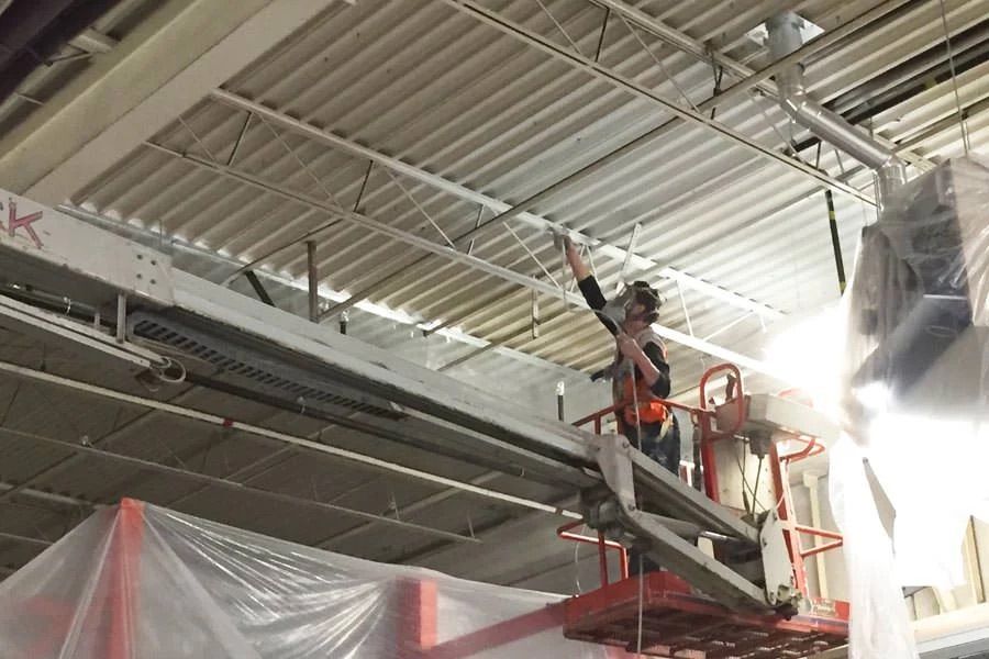 Person on a lift painting ceiling. White ceiling and steel beams. Plastic sheeting covers nearby objects.