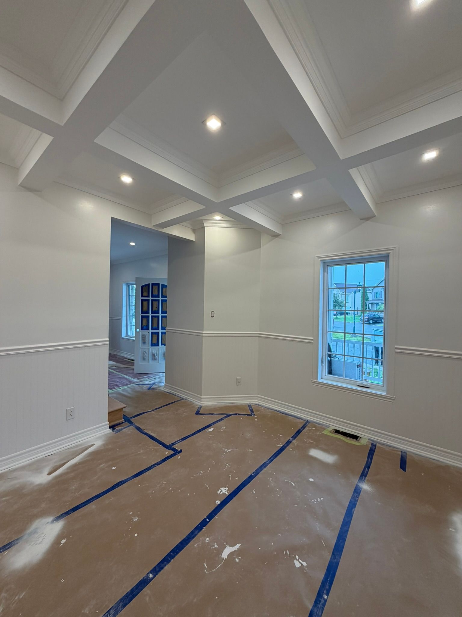 Empty room with recessed ceiling lights, taped floor protection, and windows during interior painting.