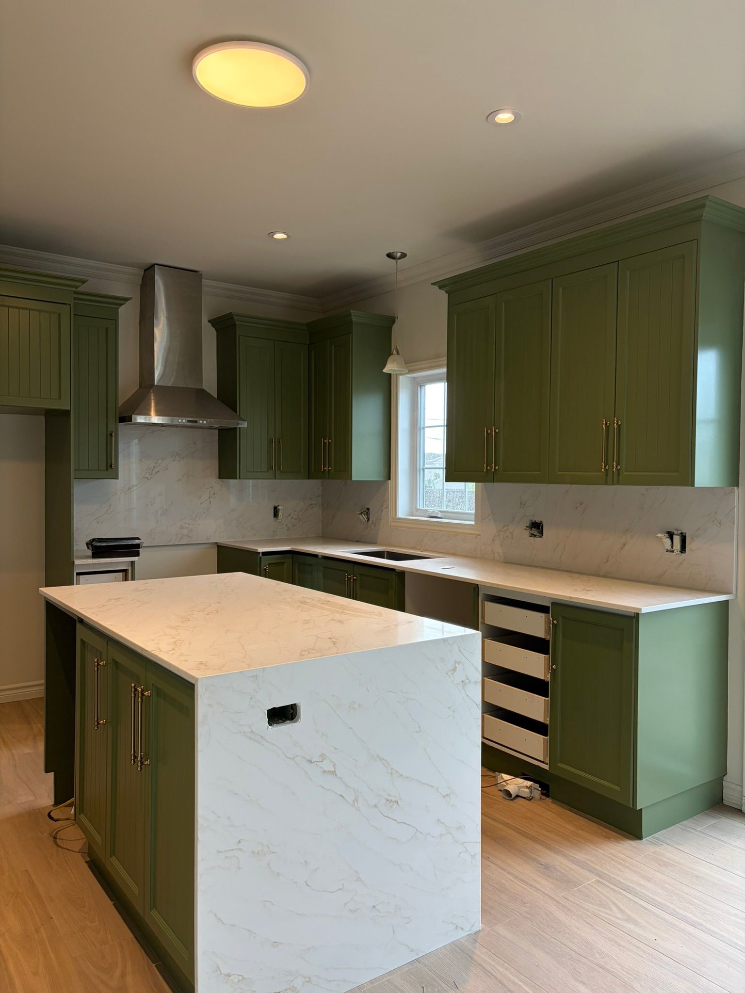 Modern kitchen with green cabinets, white marble island, stainless hood, and bright window lighting