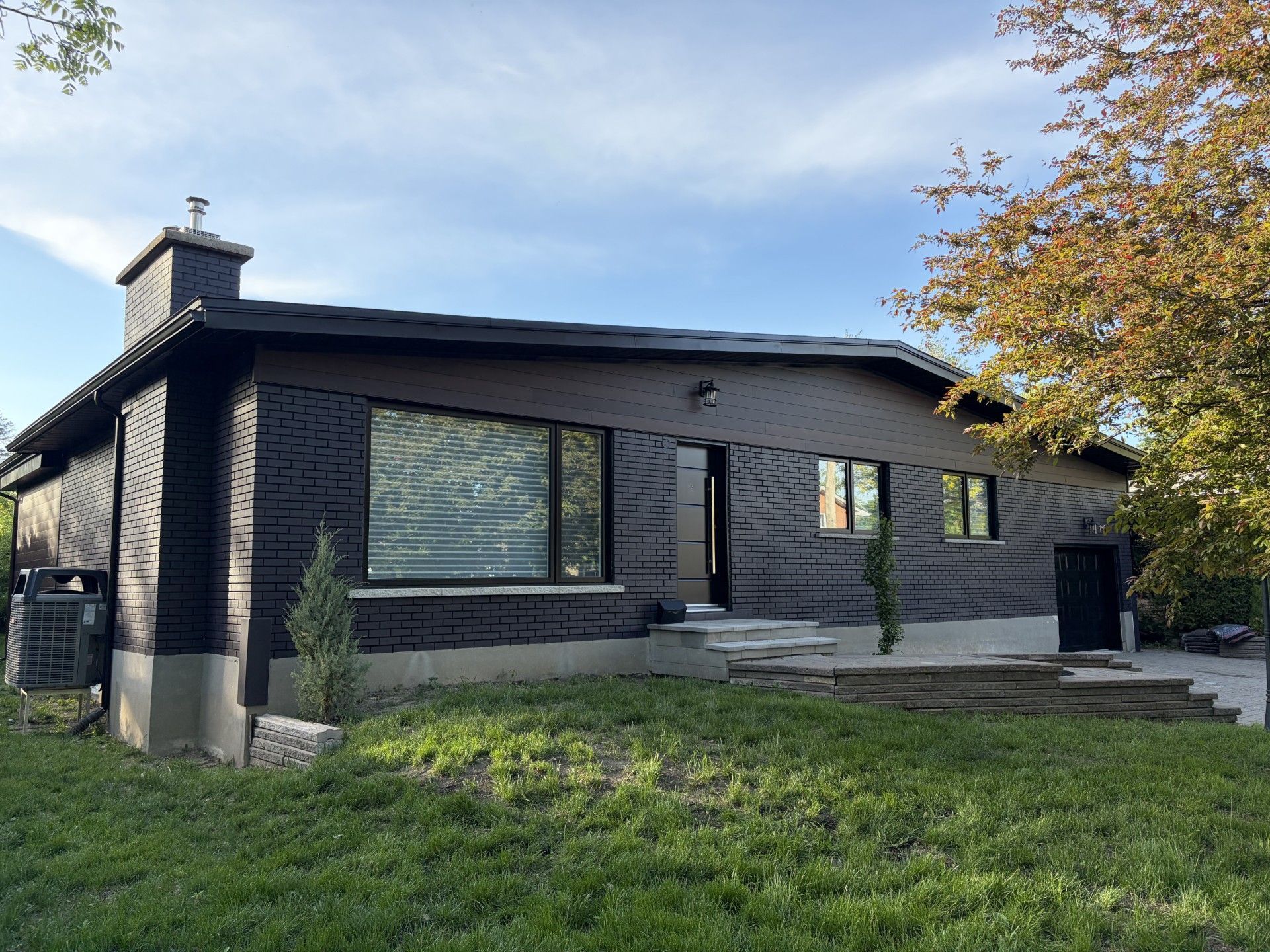 Maison moderne gris foncé avec un toit en pente, de grandes fenêtres et une cour avant gazonnée sous un ciel bleu.