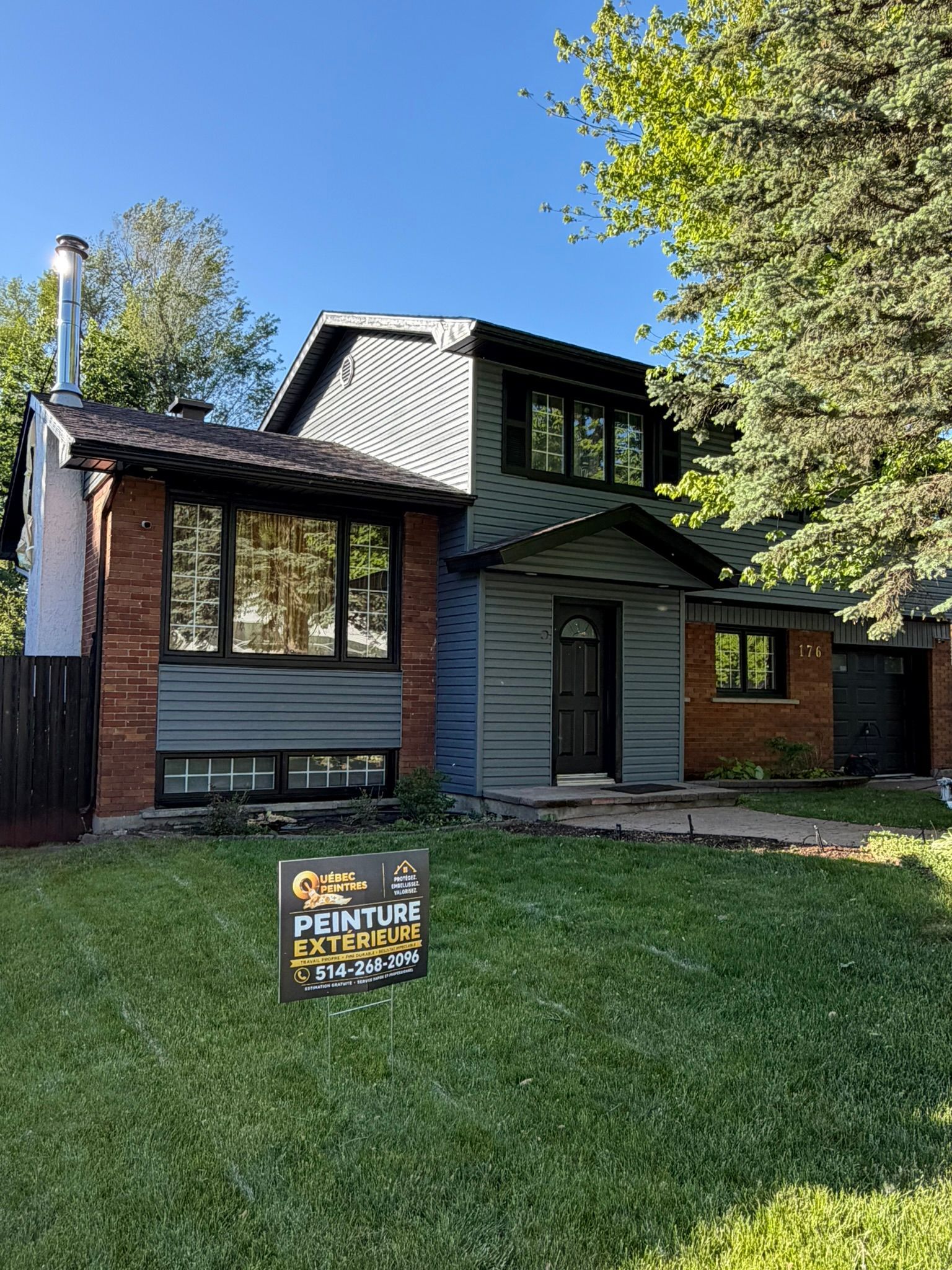 Gray two-story house with black roof and green lawn, with a small yard sign in front.