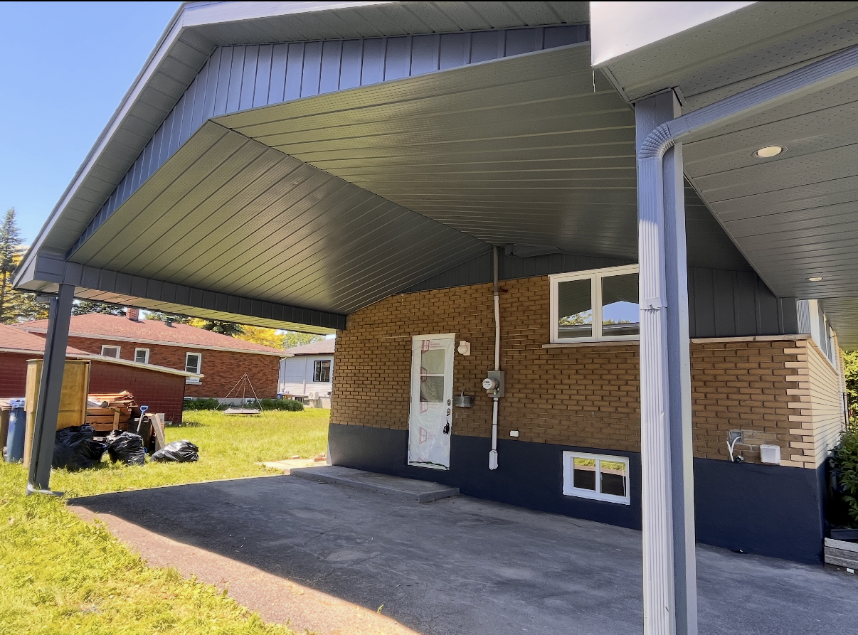 Covered patio with gray siding, white door, and brick exterior beside a grassy yard.
