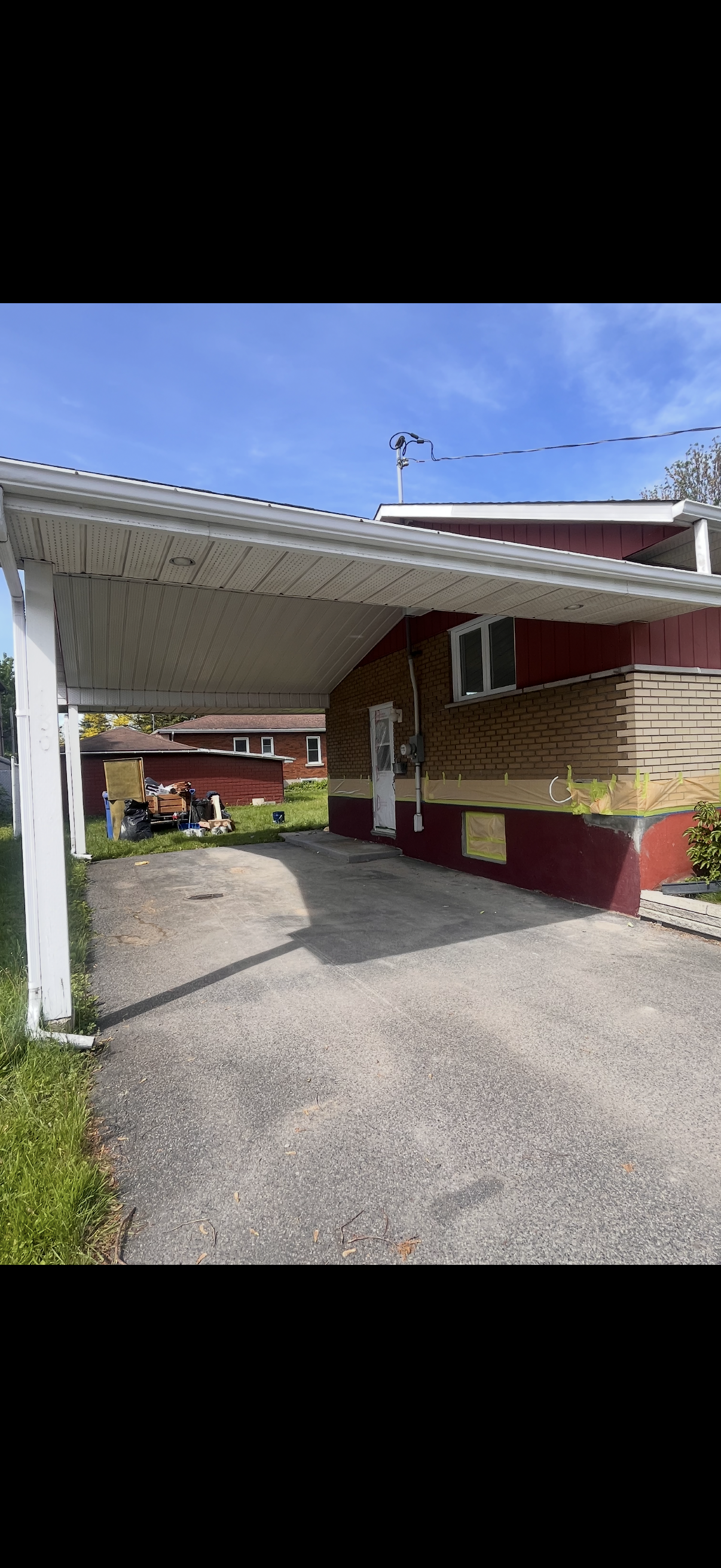 Covered driveway beside a red house with gravel ground and blue sky overhead