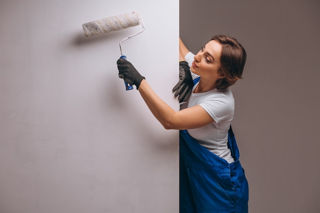 Woman in blue overalls paints a white wall with a paint roller.