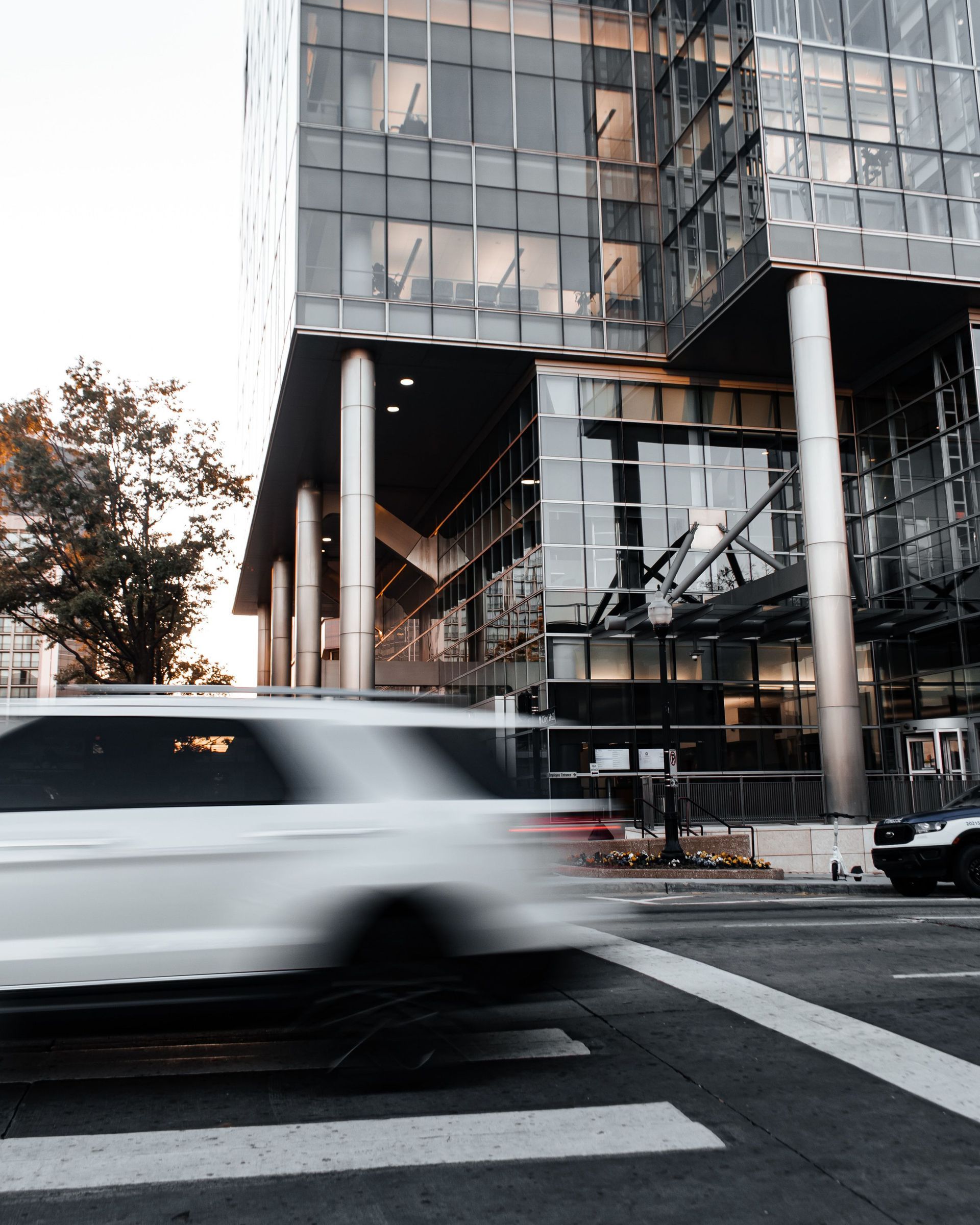 White SUV blurs past a modern glass building at a city crosswalk.