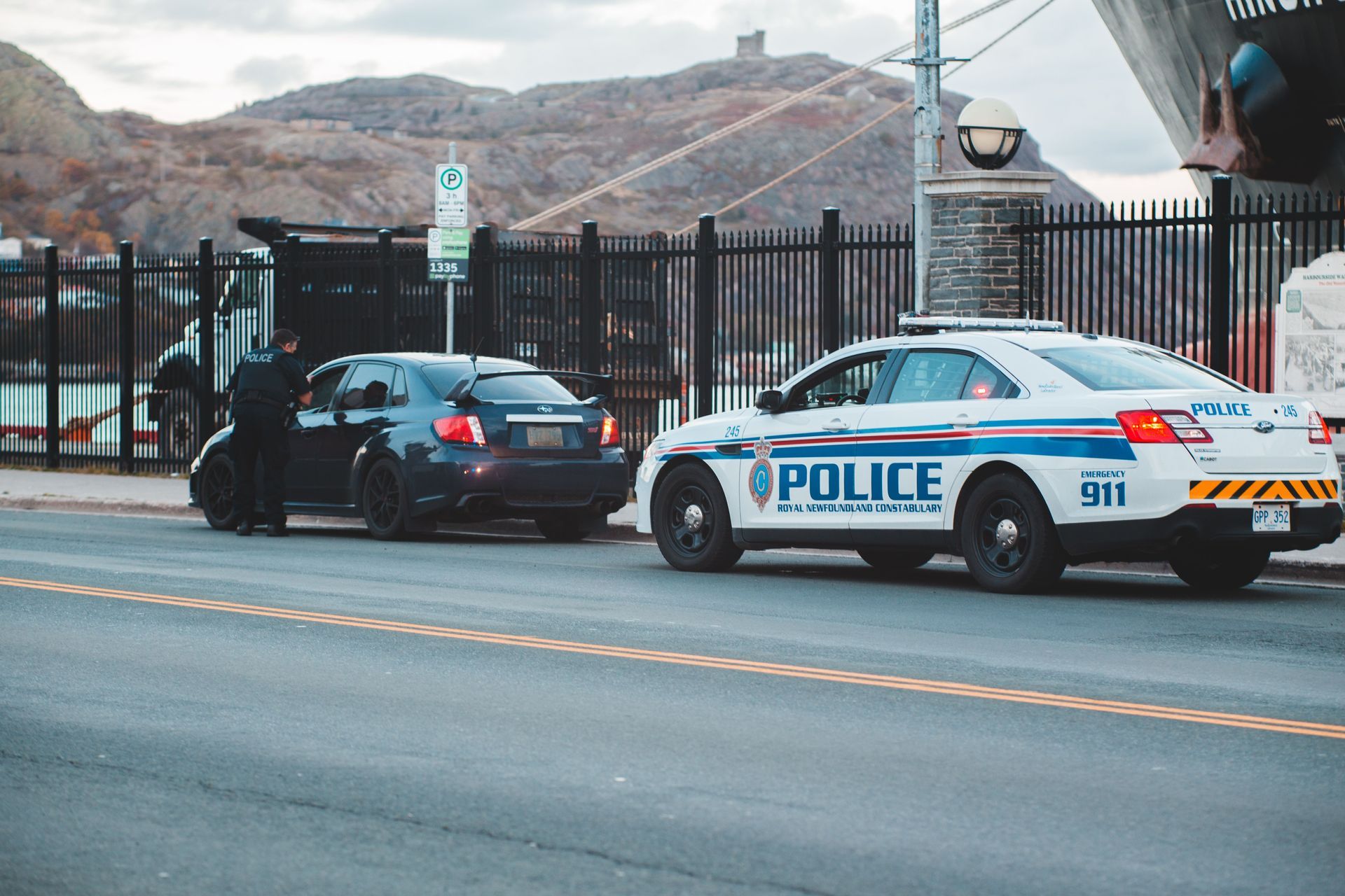 Police car and black sedan stopped on a road beside a fence, with hills in the background.