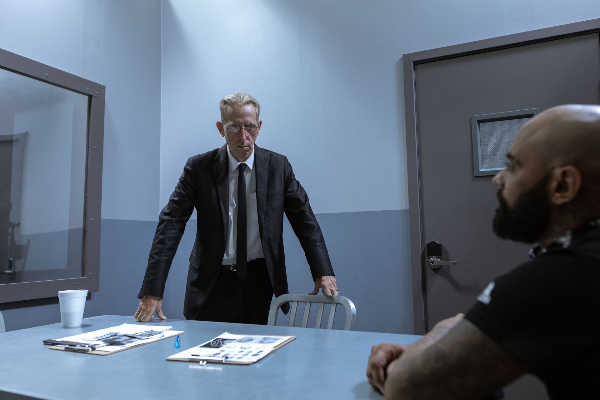 Two men face off across a table in a sparse interrogation room.