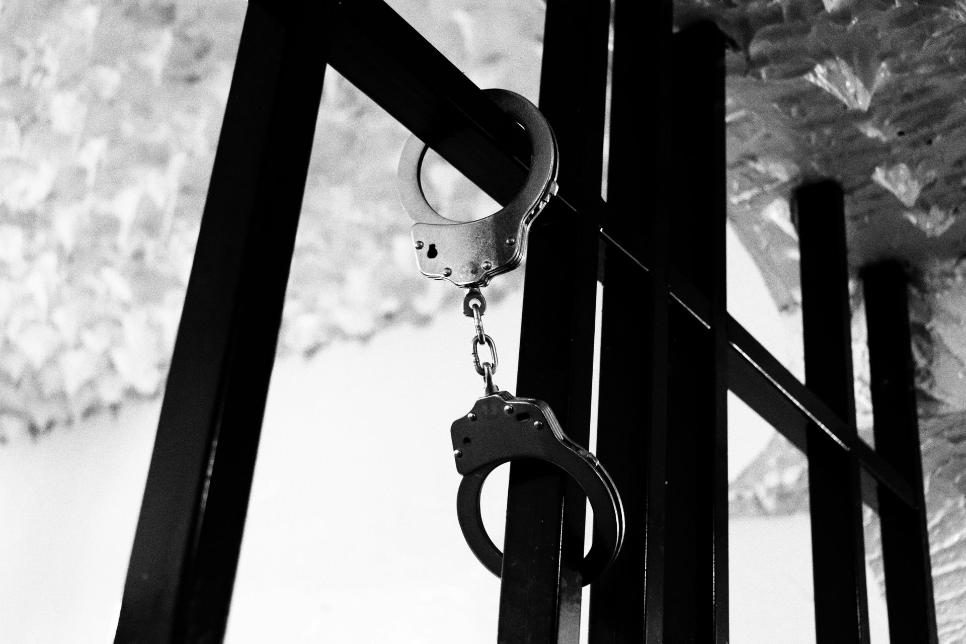 Black-and-white close-up of hanging handcuffs on metal bars with trees in the background