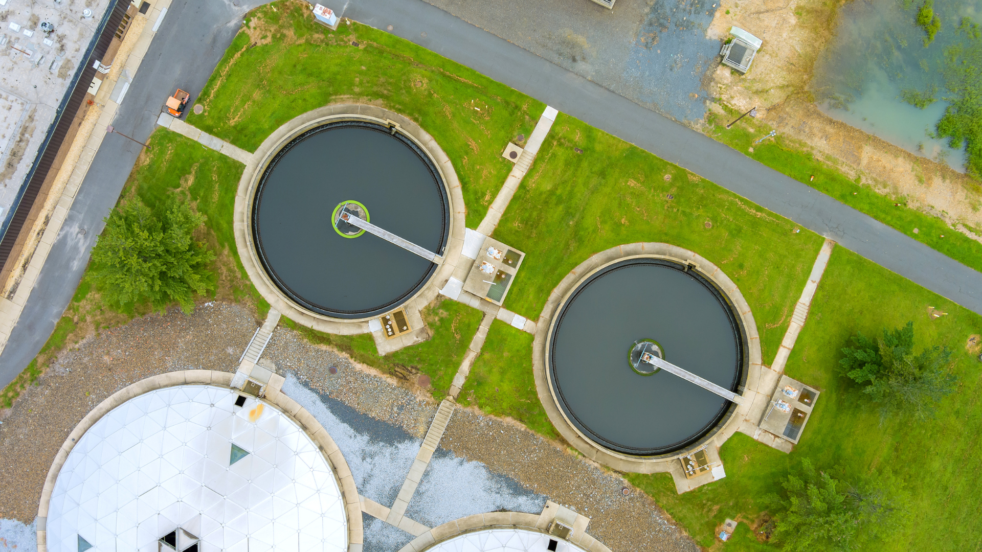 Vista aérea de una planta de tratamiento de aguas residuales con grandes tanques circulares, aguas grises y césped verde.