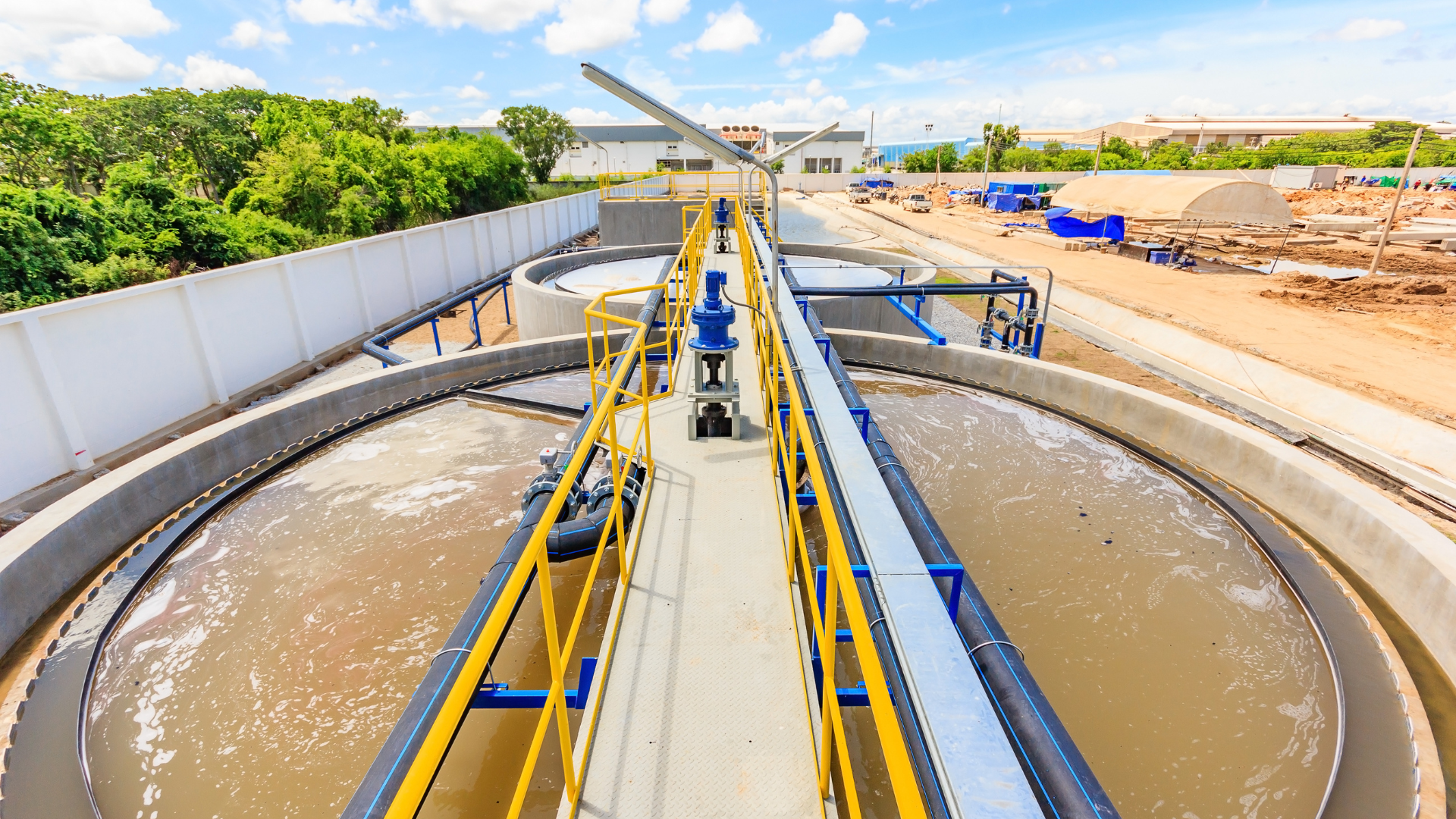 Planta de tratamiento de agua: Piletas circulares con agua marrón, pasarela metálica, estructuras azules, ambiente exterior.