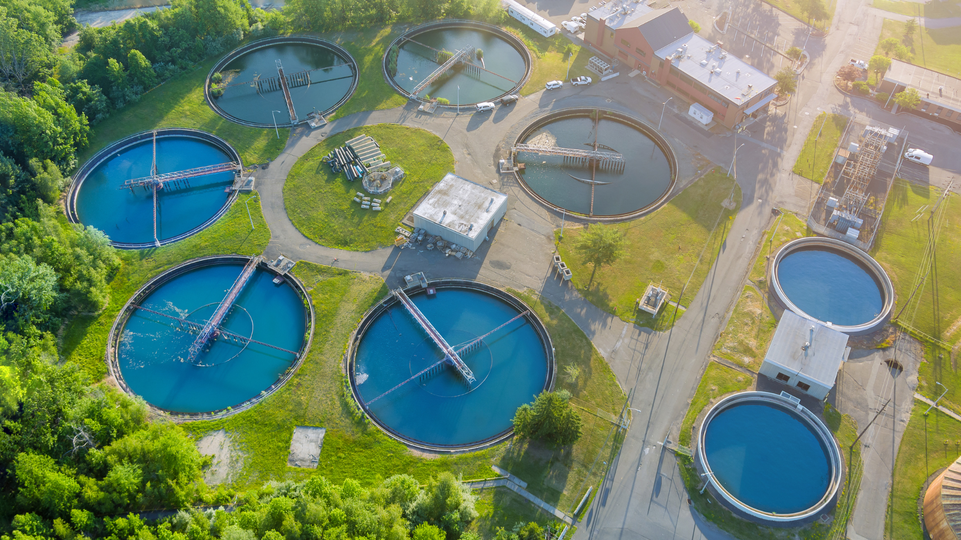 Vista aérea de una planta de tratamiento de agua con varios tanques circulares grandes. Agua azul, rodeada de césped verde y árboles.