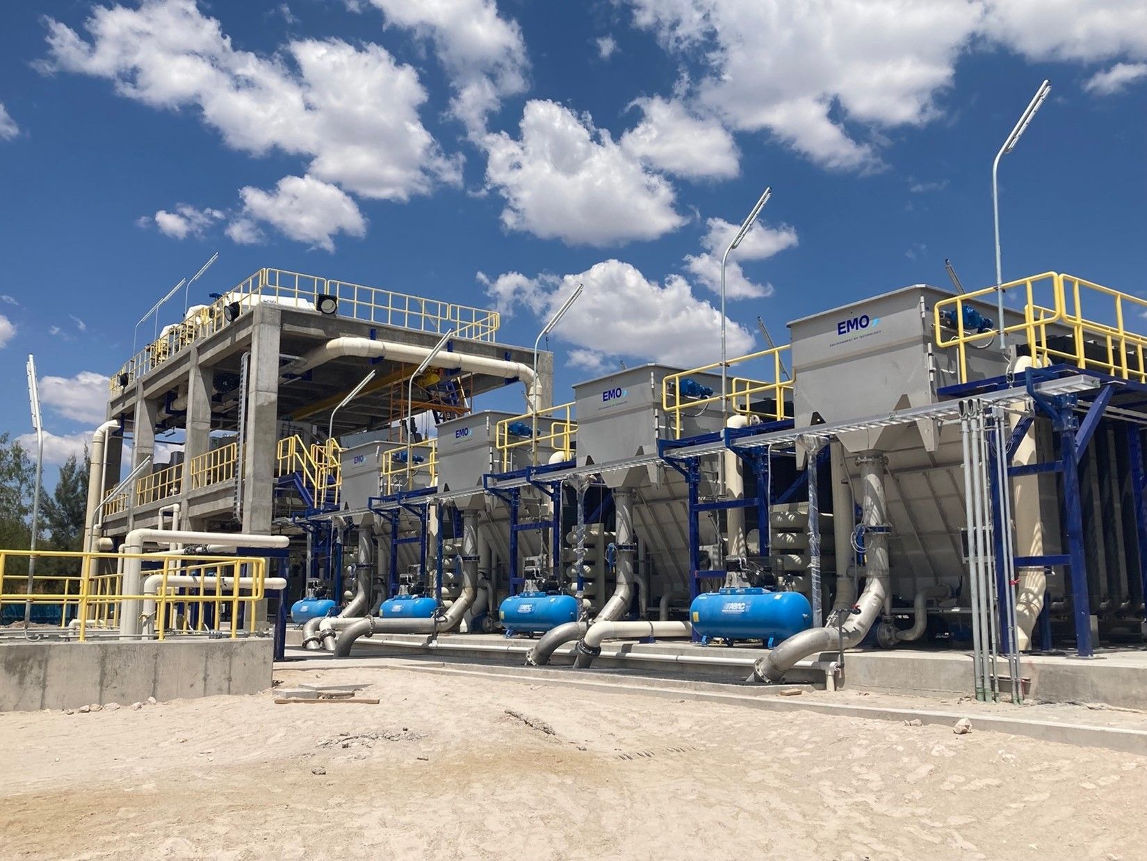 Planta de tratamiento de agua industrial bajo un cielo azul nublado. Tanques, tuberías y estructuras en tonos grises, azules y amarillos.