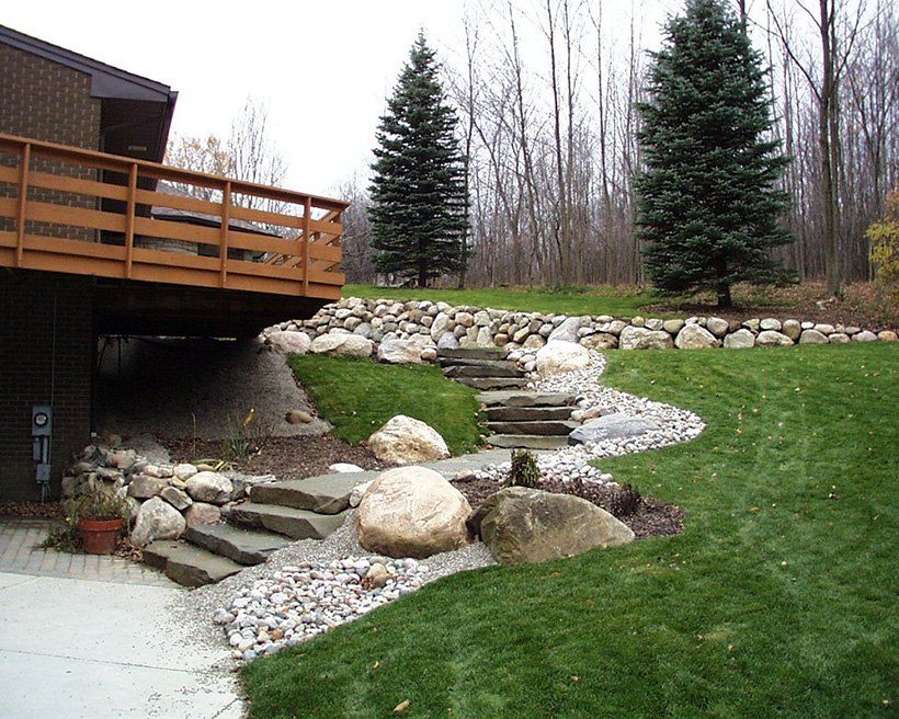 Landscaped yard with steps, grass, rocks, and trees next to a house with a wooden deck.