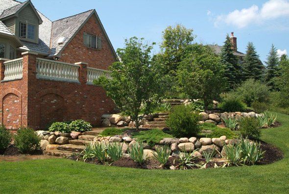 Brick house with landscaped yard featuring stone retaining walls and steps, trees, and greenery.
