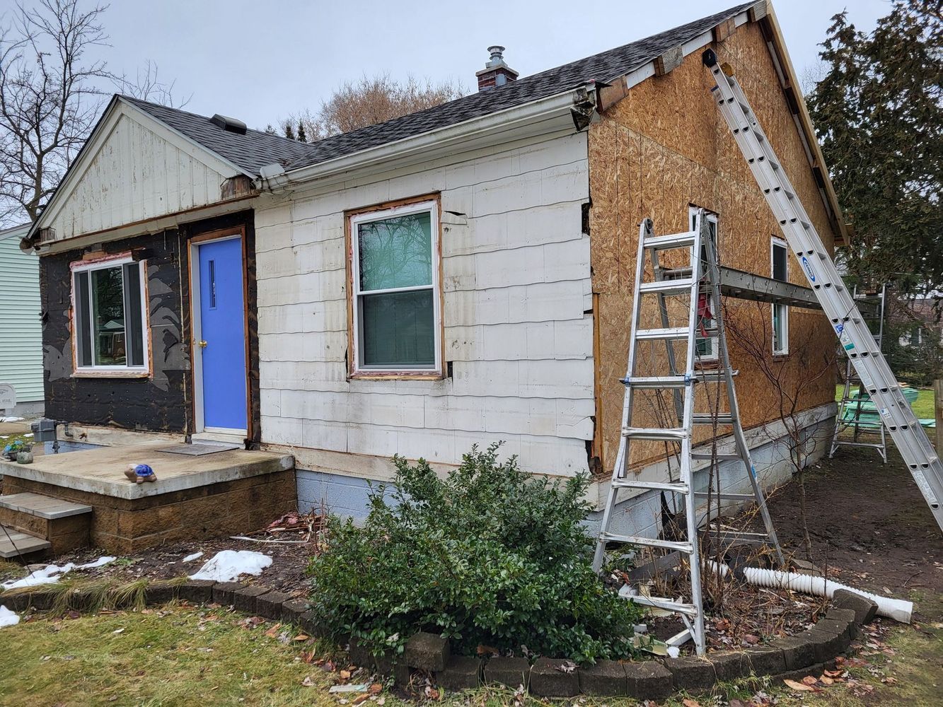 House exterior under renovation; siding removed, ladder propped, blue door, white siding, plywood exposed, steps.