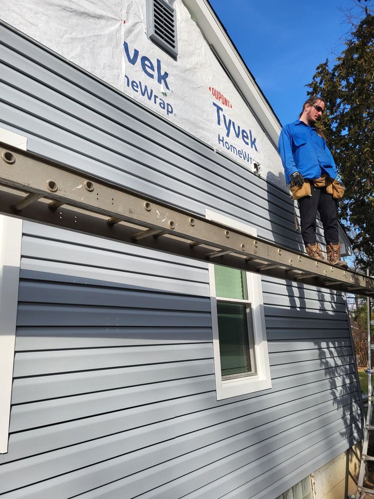 Person standing on a ladder installing gray siding on a house. Blue sky in the background.