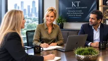 Two professionals at a desk in an office, with a laptop and notebook, appearing to discuss work
