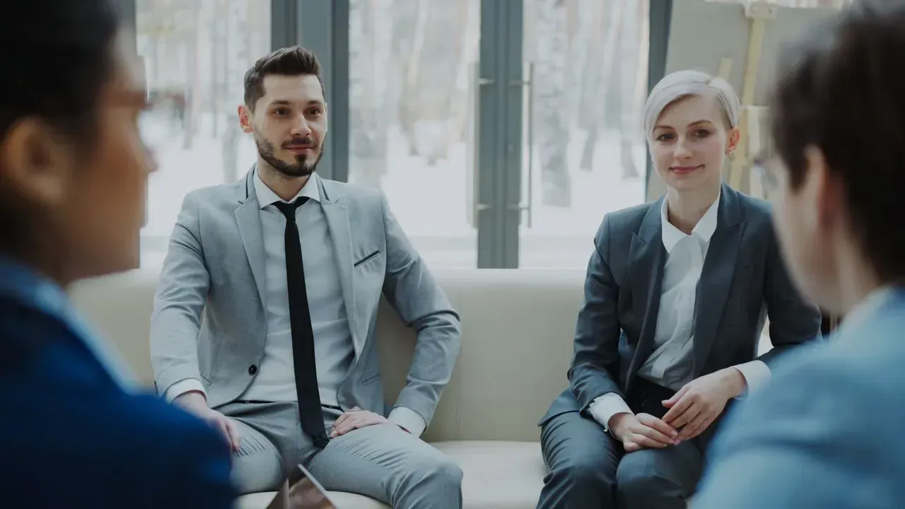 Two professionals seated in a meeting room, listening to a discussion with blurred attendees in the foreground