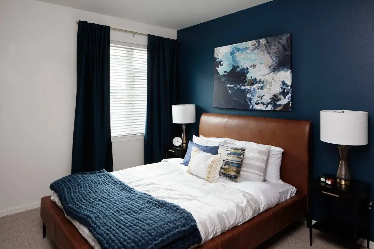 A cozy bedroom featuring a king-size bed with brown leather headboard, white linens, and a blue knit throw, against a dark blue accent wall with abstract art.