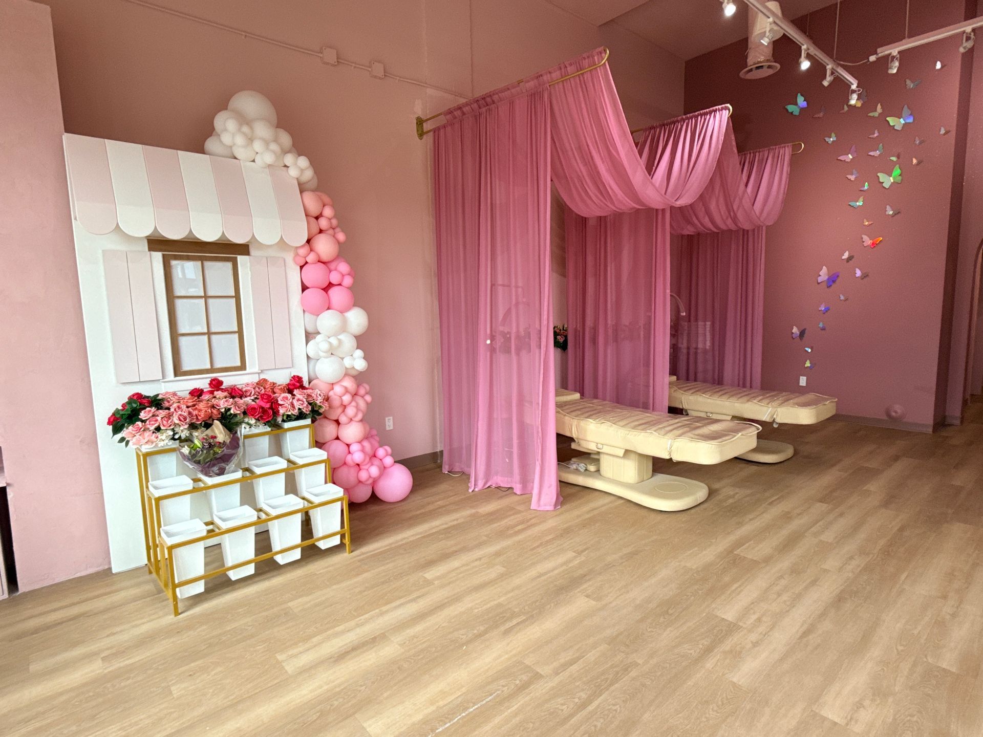 Pink party setup with canopy drapes, balloon arch, floral table, and low tables on a wood floor